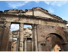 Rovine di una porta monumentale in muratura e pietra, con frontone triangolare e colonne parzialmente danneggiate, affiancata da un grande arco laterale. La struttura appare erosa e incompleta, sullo sfondo di cielo azzurro.