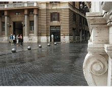 Piazza urbana lastricata con pavimentazione in sampietrini bagnati dalla pioggia, delimitata da dissuasori sferici. Sullo sfondo si vede un edificio storico in pietra con portici e vetrine, mentre in primo piano compare un elemento architettonico in pietra scolpita.