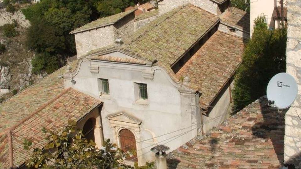 Chiesa in pietra su un pendio, con facciata semplice e tetti in coppi, affiancata da un campanile quadrato con doppie arcate. Sullo sfondo si vede una valle boscosa con alcune case sparse.
