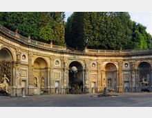 Esedra monumentale in pietra con grandi arcate e nicchie ornate da statue e rilievi, disposta a semicerchio attorno a una piazza pavimentata. Sullo sfondo si vedono alberi alti e una balaustra superiore che corre lungo la sommità della struttura.