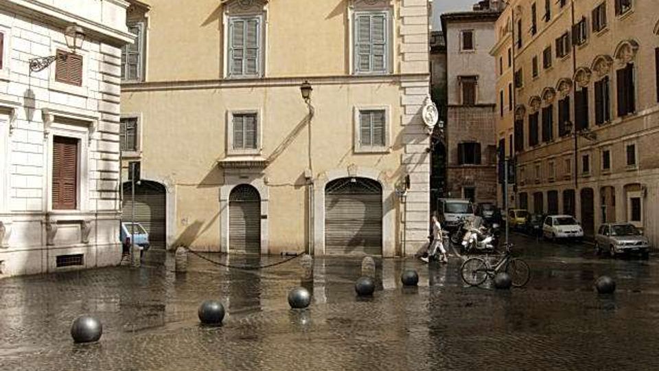 Piccola piazza urbana con pavimentazione in sampietrini bagnati e una fila di dissuasori sferici in metallo. Intorno si vedono palazzi storici intonacati con finestre e portoni ad arco al piano terra, con alcune auto parcheggiate lungo la strada laterale.
