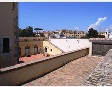 Cortile e terrazza in un complesso di edifici storici con muri intonacati gialli e bianchi, pavimentazione in mattoni e tetti in coppi. Sullo sfondo si vedono palme e altri alberi, un piccolo campanile e palazzi cittadini sotto un cielo sereno.