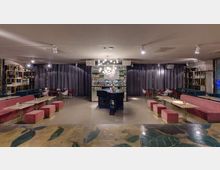 Interno di un lounge bar moderno con bancone centrale in pietra scura, lampadario a sfere luminose e scaffalature con bottiglie lungo le pareti. Ai lati sono presenti tavoli rettangolari con sedute imbottite rosa e tende scure che separano l’area del bar.