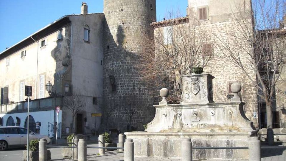 Piazza urbana con fontana in pietra al centro, decorata con stemma e tre bocchette, circondata da dissuasori. Sullo sfondo si vedono edifici in muratura e un’alta torre cilindrica in pietra, con alberi spogli ai lati.