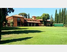 Grande edificio in mattoni con tetti in tegole rosse e ampie finestre ad arco, affacciato su un vasto prato verde. Intorno ci sono alti cipressi e altri alberi, con una struttura verde rettangolare sul lato destro.