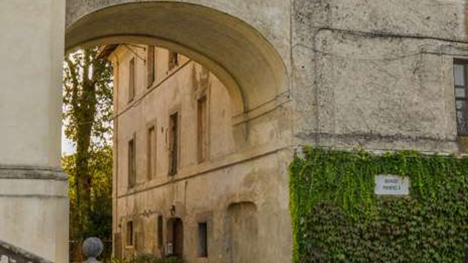 Passaggio ad arco sotto un edificio storico in muratura intonacata, con tetto in coppi e finestre a riquadri, che conduce a un cortile interno. In primo piano si vede una strada acciottolata con una breve scalinata in pietra, e sulla parete a destra un tratto coperto di edera con una piccola targa.