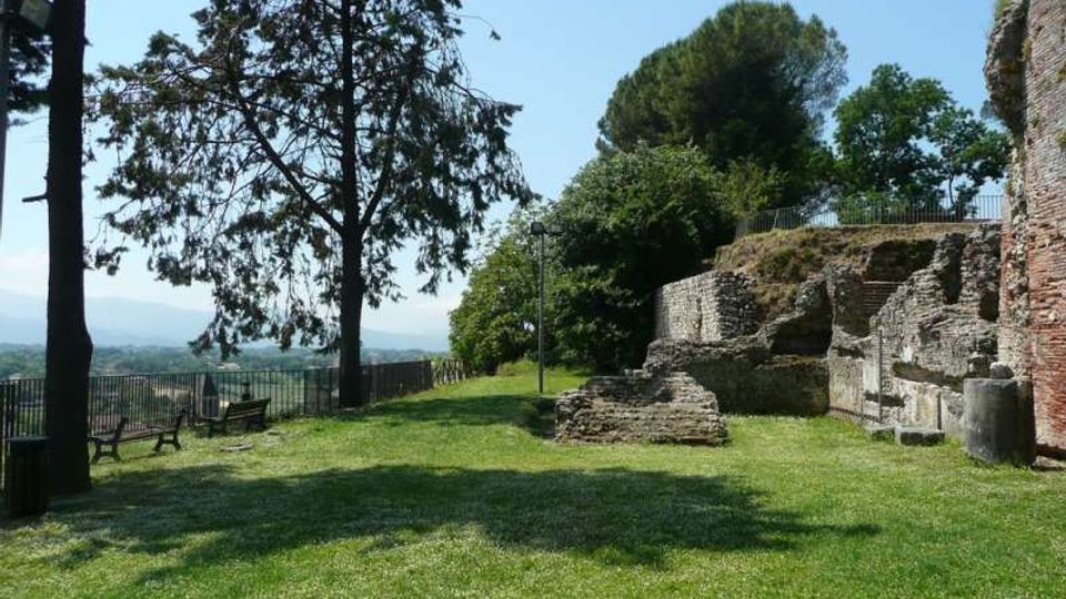 Prato alberato in un’area panoramica con panchine lungo una ringhiera, affiancato da resti di mura e strutture in pietra e mattoni. Sullo sfondo si vedono colline e un cielo sereno.
