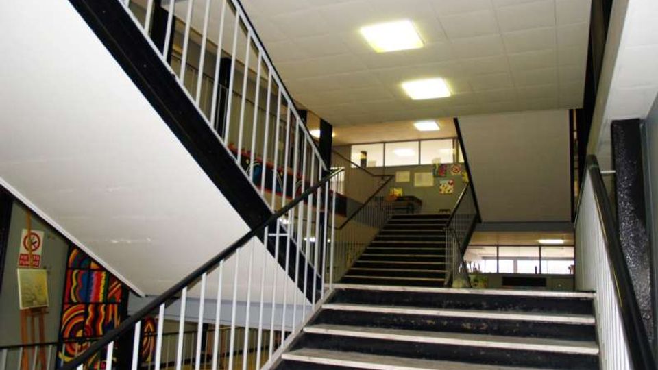 Interno di un edificio scolastico con ampia tromba delle scale: gradini scuri con bordi chiari e rampe sovrapposte con corrimano e ringhiere metalliche bianche e nere. Soffitto a pannelli con luci rettangolari e grandi finestre sul pianerottolo superiore.