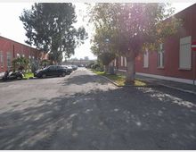 Cortile asfaltato tra due edifici bassi in mattoni rossi, con finestre a persiana e aiuole con alberi e piante lungo i lati. Sul bordo della strada sono parcheggiate diverse auto e si vede un cartello di divieto di accesso vicino all’edificio a destra.