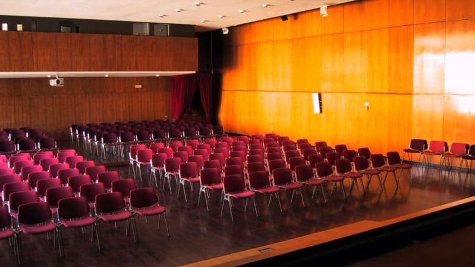 Sala auditorium con file di sedie bordeaux disposte su pavimento in legno scuro, con pareti rivestite in pannelli di legno e illuminazione a soffitto. Sul lato destro si vede un’area rialzata tipo palco e, in fondo, una tenda scura vicino all’ingresso.