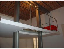 Interno di un loft su due livelli con soppalco in metallo e pavimento bianco, sostenuto da pilastri scuri e protetto da ringhiera. Soffitto alto con travi e mattoni a vista, illuminazione a spot e finestra verticale con vetri a riquadri.