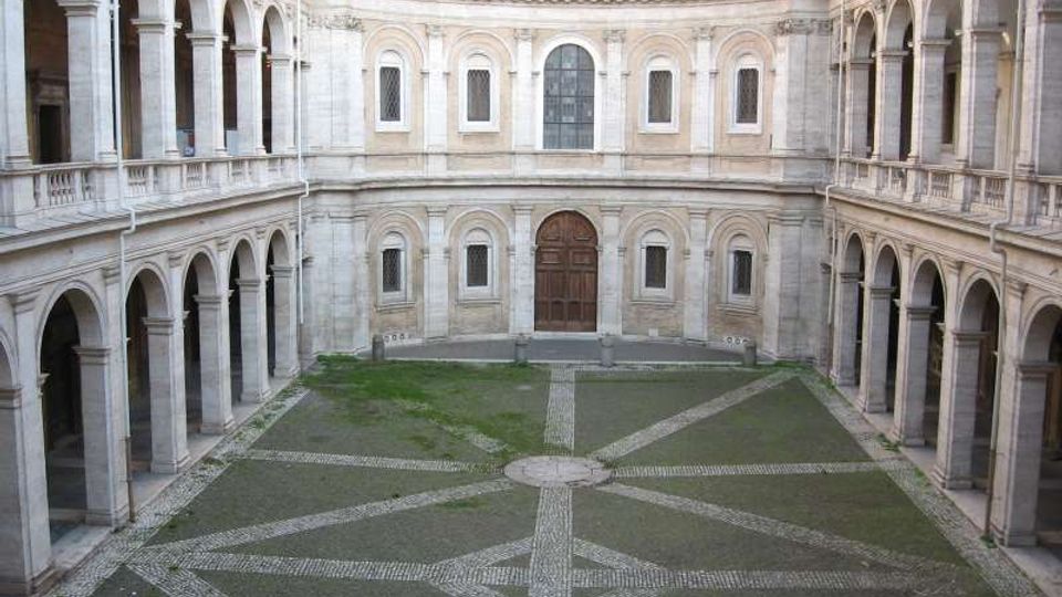 Cortile interno di un edificio storico in pietra, circondato su tre lati da logge a due piani con arcate e colonne. Al centro si vede una pavimentazione a disegno geometrico con fasce di ciottoli e un ampio portale in legno sulla parete di fondo.