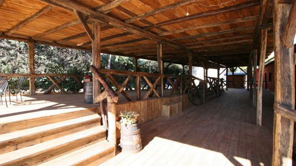 Portico rustico in legno con tetto a travi a vista e pavimento in assi, delimitato da una staccionata con montanti incrociati. A sinistra ci sono scalini e una pedana, con botti decorative e vista su alberi; il corridoio coperto si estende in profondità verso edifici sullo sfondo.