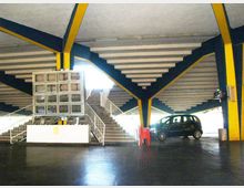 Interno di un parcheggio multipiano con ampie rampe e scale in cemento, travi strutturali verniciate in blu e giallo e pavimentazione scura. Al centro si vede una piccola area con gradinate e una struttura a finestrelle, con un’auto parcheggiata sul lato destro.