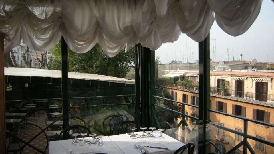 Veranda ristorante chiusa da ampie vetrate con tende bianche drappeggiate sul soffitto, tavoli apparecchiati e sedie nere in ferro. All’esterno si vede un balcone con ringhiera metallica e la vista su tetti e facciate di edifici circostanti.