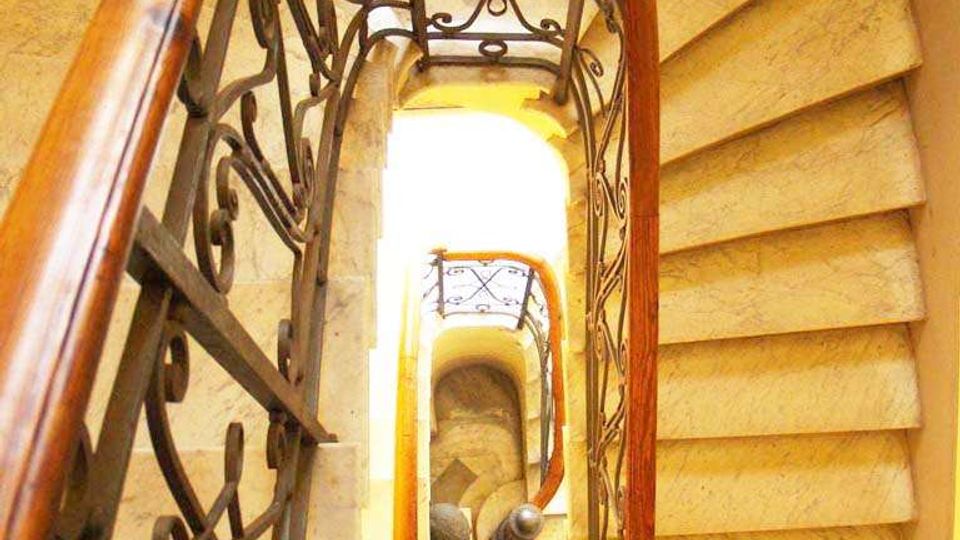 Vista dall’alto di una scala interna a chiocciola con gradini in pietra chiara, corrimano in legno e ringhiera in ferro battuto decorata. Il vano scala è illuminato da luce calda e mostra più piani che si avvitano verso il basso.