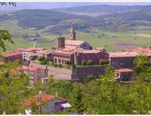 Veduta panoramica di un borgo collinare con complesso religioso in pietra e mattoni, caratterizzato da campanile e torre merlata, racchiuso da mura e circondato da case con tetti in coppi. Sullo sfondo si estendono campi coltivati e colline boscose.