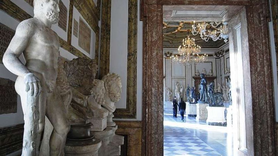 Interno di una galleria museale con statue in marmo lungo le pareti, cornici decorative e una grande apertura incorniciata in marmo che conduce a una sala ampia con pavimento a scacchi e lampadari. Sullo sfondo si vedono ulteriori sculture esposte su piedistalli in un ambiente illuminato da luce naturale.