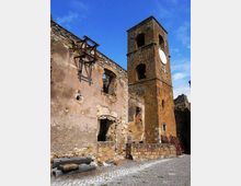 Rovine di un edificio in pietra e intonaco con muri parzialmente crollati e aperture senza infissi, accanto a una torre campanaria quadrata in pietra con grandi arcate e un quadrante circolare. La scena è in una piazzetta lastricata, con detriti e resti di strutture metalliche sotto un cielo azzurro.