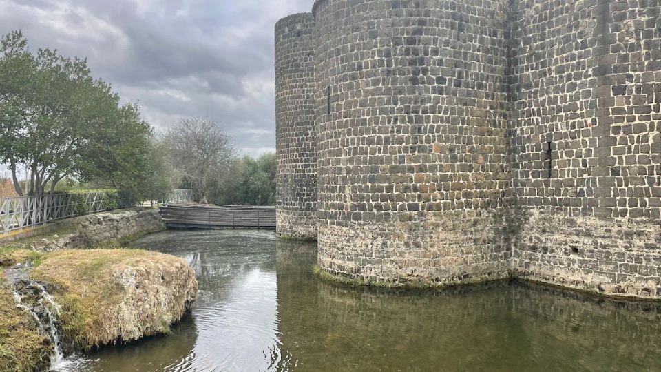 Mura di una fortificazione in pietra scura con torri cilindriche che si affacciano su un fossato d’acqua. A sinistra si vede una sponda erbosa con un piccolo salto d’acqua, alberi e una passerella in legno sotto un cielo nuvoloso.