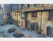 Piccola piazza lastricata in pietra con panche e tavoli in legno, circondata da case in muratura di pietra con tetti in coppi. Facciate con persiane in legno, portoni ad arco, scale esterne e numerosi vasi di fiori e piante rampicanti lungo i muri.