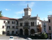 Edificio storico in pietra chiara con facciata ad archi e torre centrale merlata con orologio, affacciato su una piazza. Ai lati si vedono palazzi intonacati con tetti in tegole rosse e cielo sereno sullo sfondo.