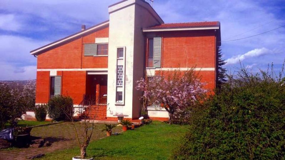 Casa unifamiliare a due piani con facciata in mattoni rossi e un alto volume centrale bianco con finestre strette verticali e ingresso con scala. Davanti si vede un giardino con prato, siepi e alberi, sotto un cielo parzialmente nuvoloso.