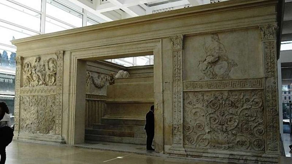 Struttura monumentale in pietra scolpita all’interno di un museo, con grande apertura centrale e pannelli decorati a bassorilievo con motivi figurativi e floreali. Ambiente moderno con pavimento lucido, ampie vetrate e soffitto a griglia metallica.