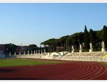 Stadio all’aperto con pista di atletica rossa che curva attorno al campo in erba e gradinate in pietra. Lungo le tribune sono allineate statue su piedistalli, con alberi alti e un edificio sullo sfondo sotto un cielo sereno.