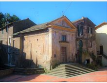 Piccola chiesa in muratura intonacata con facciata a timpano e portale centrale, affiancata da edifici in pietra. Davanti si trova una piazza lastricata con ampia scalinata che conduce all’ingresso, sotto un cielo sereno con cavi aerei visibili.