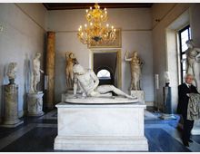 Sala museale con pavimento a scacchi e lampadario dorato, con al centro un grande basamento che sostiene una statua in marmo distesa. Lungo le pareti sono esposte diverse sculture classiche su piedistalli, con finestre alte e un passaggio ad arco sullo sfondo.
