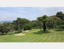 Campo da golf con ampio prato verde curato, un bunker di sabbia e alberi alti che delimitano il fairway. Sullo sfondo si vedono colline e un cielo parzialmente nuvoloso.
