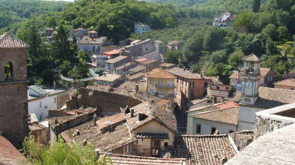 Veduta panoramica di un borgo collinare con case in pietra e tetti in coppi, stradine strette e un campanile con orologio. Il paese è circondato da colline boscose e vegetazione fitta.