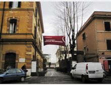 Ingresso stradale tra edifici storici in intonaco, con alberi spogli e una strada che conduce all’interno. Un grande banner rosso sopra la via indica “Musei Capitolini Centrale Montemartini”.