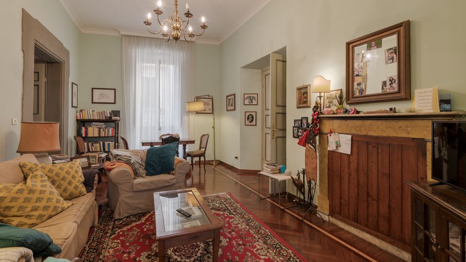 Soggiorno in stile classico con pareti verde chiaro, pavimento in parquet e grande finestra con tende bianche. Divani con cuscini, tappeto persiano e tavolino in vetro al centro, libreria e scrivania sul fondo, e camino con cornice in pietra e pannelli in legno sotto un lampadario a bracci.