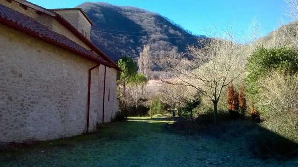 Lato esterno di un edificio in pietra con tetto in tegole rosse, affiancato da un prato e un sentiero erboso. Sullo sfondo si vedono alberi spogli e colline boscose ai piedi di una montagna sotto un cielo sereno.