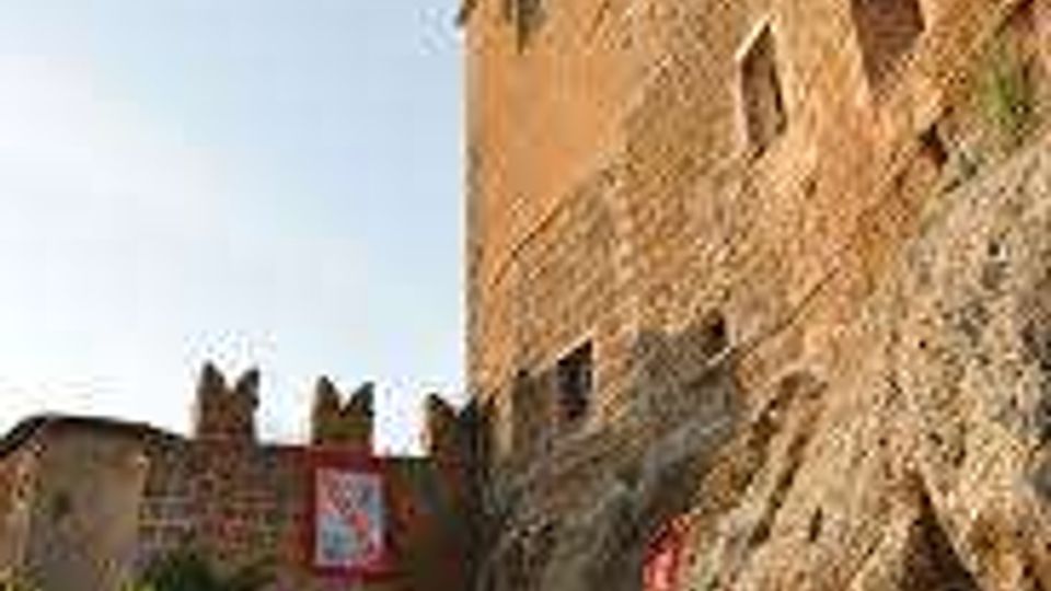 Strada in salita in un borgo medievale con arco di accesso e mura in pietra, affiancata da edifici in mattoni e una parete rocciosa a destra. Sullo sfondo si vedono torri e fortificazioni illuminate dalla luce del tardo pomeriggio.