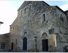 Facciata di una chiesa romanica in pietra con grandi muri spessi, archi a tutto sesto e portali scuri incassati. La struttura presenta piccole finestre ad arco nella parte alta e un tetto a due falde, affacciata su una piazza lastricata.