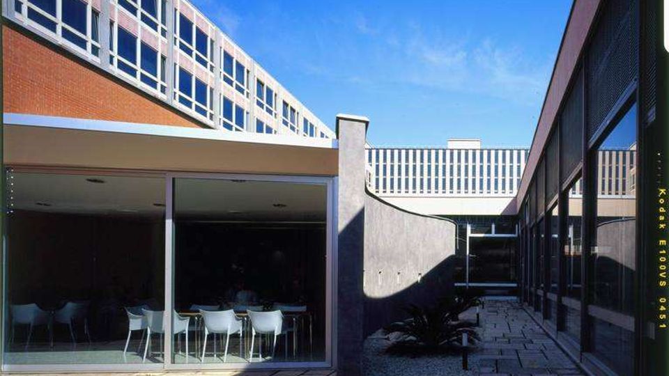 Cortile interno tra edifici moderni con facciate in mattoni e ampie finestre, collegati da un passaggio sopraelevato. Al piano terra si vede una sala con pareti vetrate e tavoli con sedie, affacciata su una pavimentazione in grandi lastre di pietra e piccole aiuole.