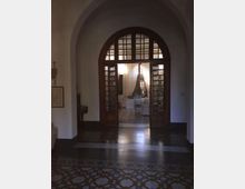 Interno di un edificio con grande arco e portale in legno a vetrate che introduce a una stanza luminosa con tende bianche e arredi chiari. Pavimento in piastrelle decorative con motivi geometrici e pareti intonacate chiare.