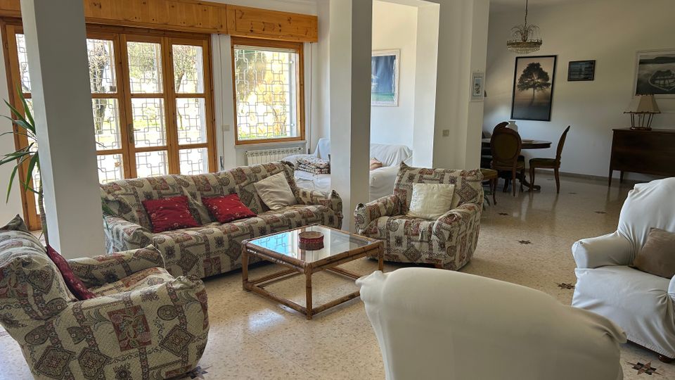 Ampio soggiorno luminoso con divani e poltrone imbottite, tavolino basso in legno e vetro e pavimento in marmo chiaro. Grandi finestre e porte-finestre con infissi in legno e grate decorative si affacciano sull’esterno, con una zona pranzo sullo sfondo sotto un lampadario.