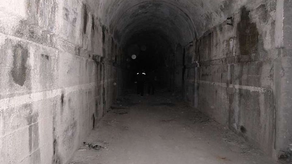 Lungo tunnel sotterraneo in cemento con soffitto a volta, pareti macchiate e pavimento polveroso. L’illuminazione è scarsa e il passaggio si perde nel buio verso il fondo.
