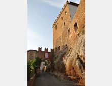 Strada in salita in un borgo medievale con arco di accesso e mura in pietra, affiancata da edifici in mattoni e una parete rocciosa a destra. Sullo sfondo si vedono torri e fortificazioni illuminate dalla luce del tardo pomeriggio.