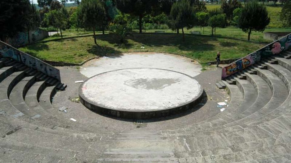 Anfiteatro all’aperto in cemento con gradinate semicircolari e un palco circolare rialzato al centro, con muri laterali decorati da graffiti. Sullo sfondo si vede un’area verde con alberi e prato.