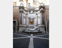 Fontana monumentale in stile barocco incassata in una parete di mattoni, con colonne laterali, timpano curvilineo e grande iscrizione centrale. In basso una vasca semicircolare con statua reclinata in marmo, su una piazzetta pavimentata a raggiera.