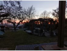 Giardino in collina al tramonto con prato, alberi spogli e un pergolato in legno con sedute e tavoli in pietra. Sullo sfondo si vedono la valle e alcune strutture lontane sotto un cielo sfumato arancione e blu.