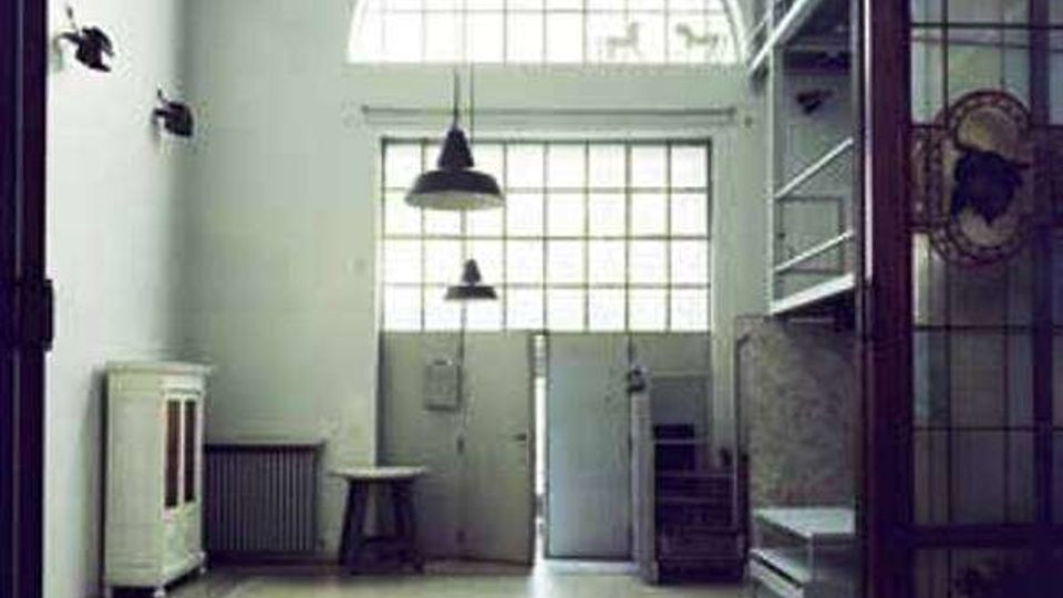Interno di un ampio spazio industriale con pavimento piastrellato, pareti chiare e grandi finestre a griglia con arco superiore sopra una porta centrale. Lampade a sospensione dal soffitto, scaffalature e una struttura a soppalco in metallo sul lato destro, con radiatore e armadietto sul lato sinistro.