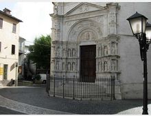 Facciata in pietra di una chiesa storica con portale ad arco riccamente scolpito e nicchie con statue, chiusa da una cancellata in ferro davanti a una scalinata. La chiesa si affaccia su una piccola piazza lastricata, con edifici bassi ai lati e un lampione in primo piano.
