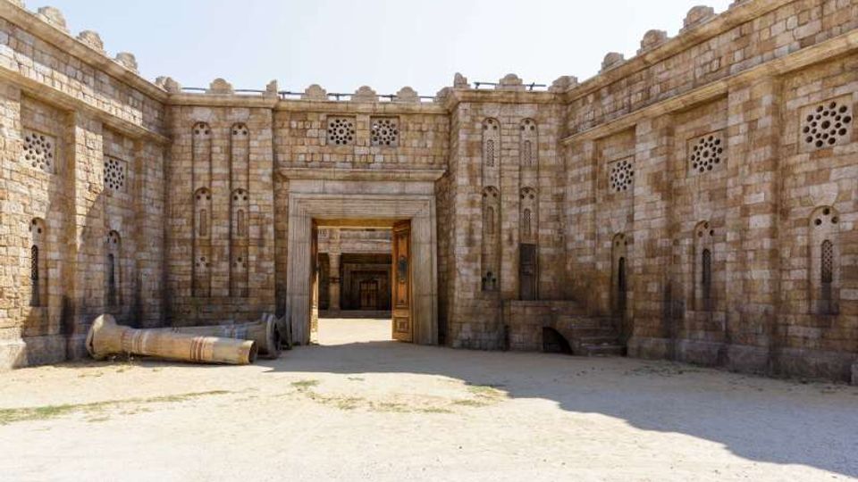 Cortile interno di una fortezza in pietra con alte mura merlate e piccole finestre ad arco e traforate. Al centro si apre un grande portale rettangolare che conduce a un’area ombreggiata; sul terreno sabbioso è visibile una colonna di pietra caduta.