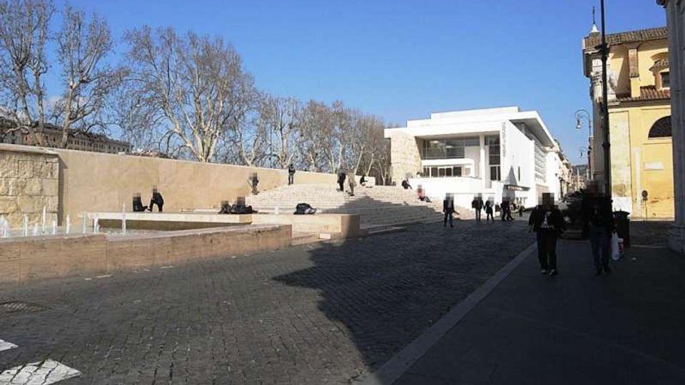 Piazza urbana con ampia scalinata in pietra e piccola fontana a raso sul lato sinistro, affacciata su un edificio moderno bianco con grandi vetrate. Sullo sfondo ci sono alberi spogli e lampioni lungo il marciapiede accanto a edifici storici.